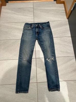 Levis 512 - Säljer ett par blå jeans med raka ben och slitningar på båda benen. Jeansen har klassisk femficksdesign och knappar i metall. Materialet är denim och färgen är en tvättad blå nyans med ljusare partier. Priset står inte skrivet i sten! Hör av er för minsta fundering!