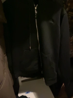 Svart hoodie från Burberry med dragkedja - Snygg svart hoodie från Burberry med dragkedja framtill och klassiskt rutigt foder i huvan. Hoodien har justerbara snören vid huvan och fickor på sidorna. Perfekt för dig som vill ha en stilren och exklusiv look. Kommer i själva burberry påsen med.