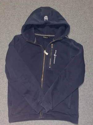 Marinblå hoodie från Sail Racing - Snygg marinblå hoodie från Sail Racing med dragkedja och broderad logga på huvan. Hoodien har två fickor framtill och vit Sail Racing-tryck på bröstet. Tillverkad i mjukt bomullsmaterial och har sportig känsla med randig detalj i kragen. Storlek 170