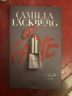 Vingar av silver av Camilla Läckberg - Spännande roman av Camilla Läckberg med titeln 'Vingar av silver'. Omslaget är grått med röd text och ett läppstift i mitten. Boken handlar om Faye och hennes kamp för hämnd och överlevnad. Perfekt för dig som gillar thrillers.