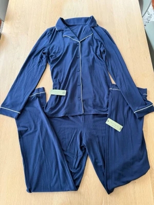 Mörkblå pyjamas med vita detaljer - Snygg mörkblå pyjamas från Intimissimi med långärmad skjorta och byxor. Setet har vita kontrastsömmar längs kanterna och knappar framtill. Skjortan är i storlek S och byxorna i M.💕Pjamasen är oanvänd och lapparna är ännu fast!