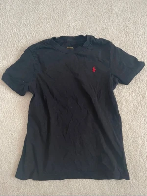 Svart t-shirt från Polo Ralph Lauren - Svart t-shirt från Polo Ralph Lauren med klassisk passform och korta ärmar. T-shirten har en liten röd broderad logga på bröstet. För barn i storlek 6-7 år gammal!!