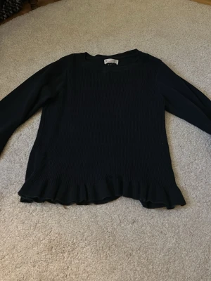 Svart blus med volangkant - Stilren svart blus med långärmad design och volangkant nedtill. Blusen har en smockad framsida som ger en snygg struktur och är tillverkad i ett lätt, mjukt tyg. Perfekt att matcha med jeans eller kjol för en trendig look.