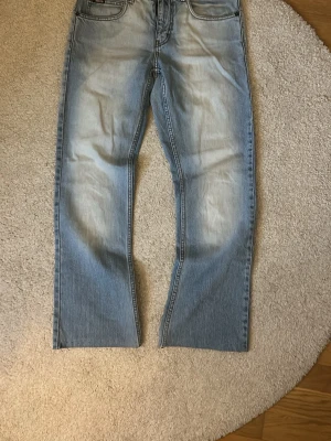 Ljusblå raka jeans från Nudie - Säljer ett par ljusblå jeans från Nudie med raka ben och klassisk femficksmodell. Jeansen har snygga slitningar och kontrastsömmar, samt Nudie-logga på bakfickan. Tillverkade i bomull för en skön och avslappnad look.