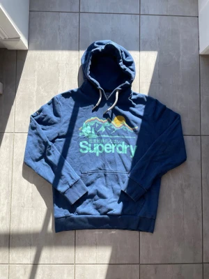 Blå hoodie från Superdry - Snygg blå hoodie från Superdry med färgglatt bergsmotiv och sol på bröstet samt logga. Klassisk känguruficka framtill och vita dragsnören i huvan. Perfekt för chill dagar och streetwear-stil. Mjuk bomullskänsla och ribbade muddar vid ärmar och midja. Passar strl: M-L, vid frågor eller funderingar skriv gärna!