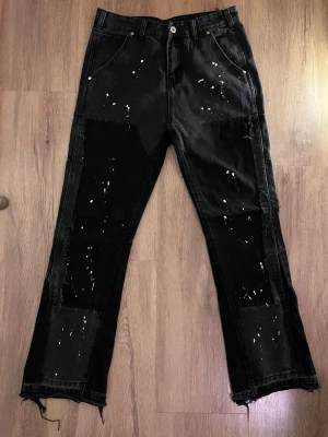 Svarta flared jeans med färgdetaljer – trendiga & unika - Säljer ett par svarta jeans med vita färgstänk och patchade detaljer på benen. Jeansen har raka ben, råa och fransiga benslut samt flera fickor både fram och bak. Materialet är denim och de har en avslappnad, street-inspirerad look.