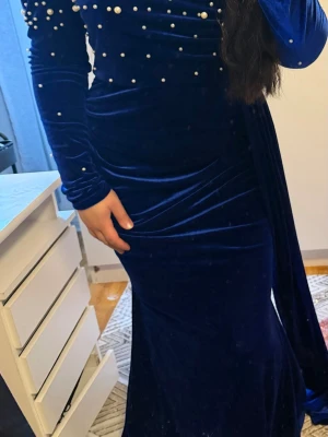 Mörkblå sammetsklänning med pärlor - Elegant långklänning i mörkblå sammet med långa ärmar och dekorativa pärlor över bröstet och axlarna. Klänningen har figurnära passform och en lyxig känsla, perfekt för speciella tillfällen. Snygg drapering och klassisk siluett.