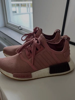 Adidas NMD R1 rosa sneakers - Säljer ett par rosa Adidas NMD R1 sneakers med stickad ovandel, klassiska tre ränder på sidan och vit Boost-sula. Skorna har snörning och detaljer i mörkare rosa samt svarta block på sulan. Perfekta för en streetwear-look.