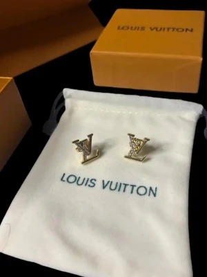 💛 Louis Vuitton LV örhängen guld - Snygga örhängen från Louis Vuitton i guldfärgad metall med ikoniska LV-loggan. Ena örhänget är dekorerat med små gnistrande stenar för extra bling. Kommer med originalpåse och ask, perfekt för dig som gillar lyxiga accessoarer.
