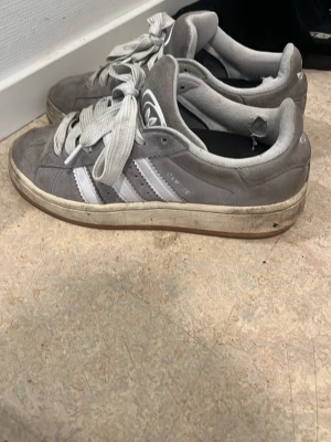 Skor o topp  - Ett par grå sneakers från Adidas med klassiska vita ränder på sidan och vita skosnören. Skorna har en låg profil och är tillverkade i mocka med gummisula. Perfekt för dig som gillar sportig och avslappnad stil.