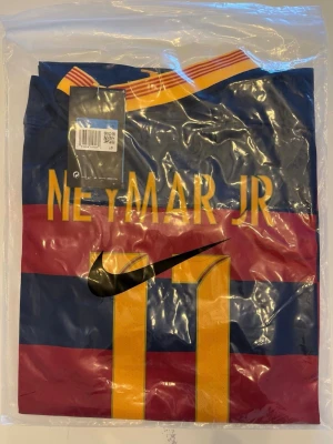 Barcelona Neymar JR matchtröja - Säljer en helt ny Barcelona matchtröja med Neymar JR och nummer 11 på ryggen. Tröjan har breda blå och röda ränder, gula detaljer och Nike-logga. Perfekt för dig som älskar fotboll och vill ha en ikonisk supportertröja.