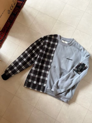 Grå och rutig sweatshirt Unknown - Unik sweatshirt i grått med svart, vitt och rött rutigt tyg på ena sidan och ena ärmen. Tryck på bröstet och ryggen samt rutigt tygdetalj på ena ärmen. Mjuk bomullsmix och avslappnad passform. Perfekt för en cool streetwear-look.