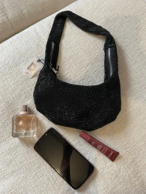 Svart glittrig handväska från Mango - Cool svart handväska från bikbok med glittriga pärlor över hela väskan. Väskan har en bred axelrem och två silverfärgade dragkedjor på sidorna. Perfekt för dig som vill sticka ut och addera lite extra bling till din outfit.
