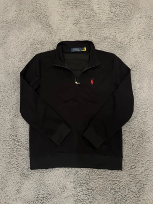 Svart halvzip från Polo Ralph Lauren - Svart långärmad tröja från Polo Ralph Lauren med halv dragkedja och klassisk röd broderad logga på bröstet. Tröjan har ribbade muddar och krage, perfekt för en stilren och avslappnad look. ⚠️tröjan har bara använts 2 gånger⚠️