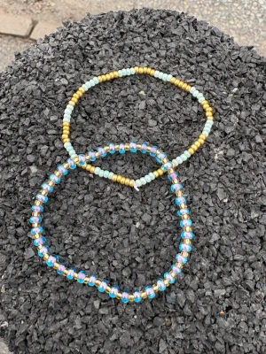 Två färgglada pärlarmband - Säljer två handgjorda armband med små pärlor. Ett armband har turkosa och guldiga pärlor, det andra har skimrande blå, lila och guldiga pärlor. Perfekta för att ge din outfit en färgklick. Elastisk tråd gör dem enkla att ta på och av.