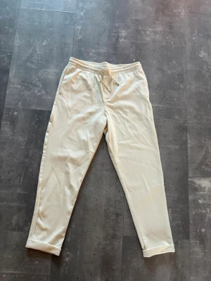 Beige mjukisbyxor - Säljer ett par stilrena beige mjukisbyxor med resår i midjan och dragsko för skön passform. Byxorna har raka ben och uppvikta benslut, samt fickor på sidorna och baktill. Perfekta för chill dagar eller när du vill vara bekväm.