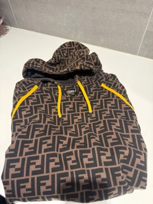 Fendi brun hoodie med FF-mönster - Säljer en brun hoodie från Fendi med ikoniskt svart FF-mönster över hela tröjan. Hoodien har gula dragsnören och en klassisk huva. Perfekt för dig som gillar lyxiga streetwear-plagg med tydlig logga. Endast tvättad på kemtvätt, enda synen på användning är den lätta blekningen på midjemuffen som syns på sista bilden. Nypris ungefär 10000kr
