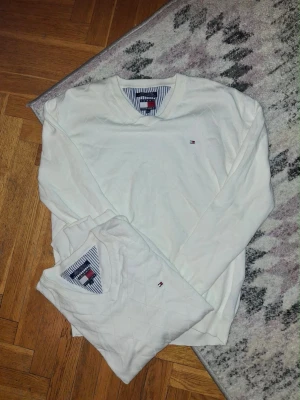 Vit tröja Tommy Hilfiger XL - Klassisk vit tröja från Tommy Hilfiger i 100% bomull. Tröjan har V-ringning, diskret logga broderad på bröstet och ribbade muddar. Perfekt för en clean och stilren look.