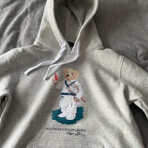 Grå hoodie Polo Bear Ralph Lauren - Säljer en ljusgrå hoodie från Ralph Lauren med den ikoniska Polo Bear-trycket framtill. Hoodien har huva med vita snören och en stor magficka. Materialet är mjukt och skönt, perfekt för en avslappnad stil. Klassisk och trendig design som alltid funkar. Små i storleken så S funkar också. Mycket bra skick! Hör av er vid funderingar! (Pris går att diskutera vid snabb äffär!)