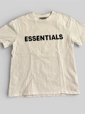 Vit Essentials t-shirt med tryck - Vit t-shirt från Essentials med svart texttryck på bröstet. Större passform som passar både S och M med rund halsringning och korta ärmar. Tillverkad i mjuk bomull som känns skön mot huden. Perfekt för en avslappnad och stilren look. 