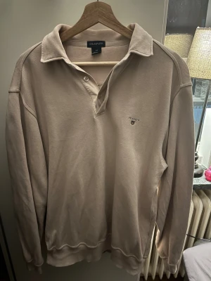 Gant Beige långärmad  - Klassisk beige långärmad tröja från GANT med krage och knappslå. Tröjan har broderad logotyp på bröstet och är tillverkad i ett mjukt, strukturerat material. Perfekt för en stilren och avslappnad look.