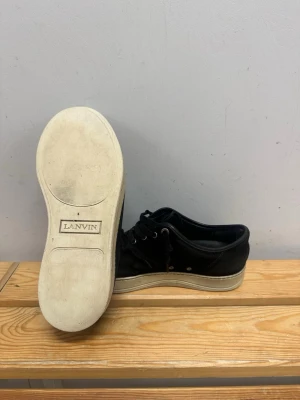 Svarta Lanvin sneakers i mocka och läder - Stilrena svarta sneakers från Lanvin med ovandel i mocka och läder, vita gummisulor och svarta skosnören. Klassisk låg modell med metallöljetter och diskret design som passar till det mesta. Och men får box också 