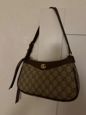 Gucci ophidia väska - Säljer en snygg och oanvänd axelväska. Väskan är beige med bruna detaljer i läder, guldfärgat GG-spänne och justerbar axelrem. Väskan är en AA kopia och ser nästintill helt äkta ut. Köpt för 1500 kronor. Har gjort ett eget hål i axelremmen för att den ska komma till den storleken som den gör på bilden. Orginal längden finns på sista sliden. Pris kan diskuteras. 