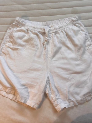 Vita linne shorts med snörning från H&M - Säljer ett par vita shorts från H&M med relaxed fit och snörning i midjan. De har elastisk midja och två sidofickor. Perfekta för varma dagar och enkel att matcha med olika toppar.