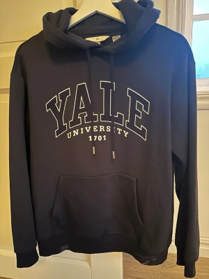 Svart Yale hoodie från H&M - Snygg svart hoodie från H&M med stor vit YALE UNIVERSITY-print på bröstet och känguruficka framtill. Klassisk huva med mjukt material som känns skönt mot huden. Perfekt för dig som gillar college-stil och vill ha en avslappnad look.