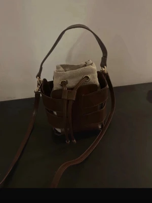 Brun bucket väska - Snygg brun bucket handväska med flätad design och guldfärgade detaljer. Väskan har både handtag och axelrem samt en tygpåse inuti som stängs med snörning. Perfekt för dig som vill ha en unik och trendig accessoar.