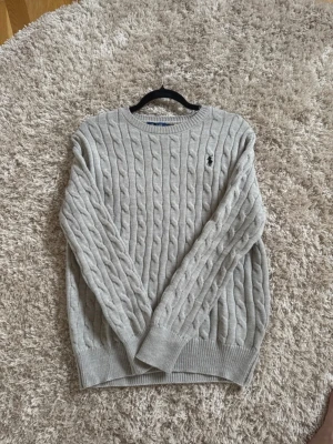  Polo Ralph Lauren tröja  - Säljer en ljusgrå kabelstickad tröja från Polo Ralph Lauren med klassisk rund halsringning och svart broderad logga på bröstet. Tröjan har ribbade muddar vid ärmslut och nederkant och är tillverkad i mjukt stickat material.