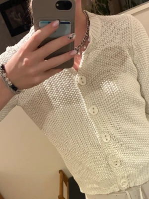 Vit stickad kofta med stora knappar - Snygg vit stickad kofta med stora matchande knappar framtill och ribbad krage. Koftan har korta ärmar och är gjord i ett grovt stickat material som ger en cool struktur. Perfekt att slänga över en t-shirt för en chill look.