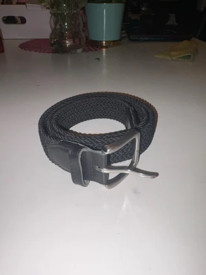 Svart flätat bälte med silverfärgat spänne - Snyggt svart flätat bälte med ett stilrent silverfärgat metallspänne. Bältet har detaljer i skinn vid ändarna och passar perfekt till jeans eller chinos. Enkel att justera och ger en clean look till outfiten.