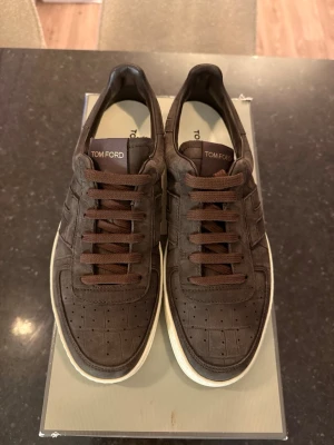 Tom Ford skor  - Snygga bruna sneakers från Tom Ford i mocka med perforerade detaljer och matchande bruna skosnören. Skorna har en vit sula och diskret logga på plösen. Perfekt för dig som gillar stilrena och exklusiva sneakers med lyxig känsla.