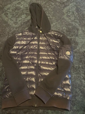 Mörkblå Moncler cardigan  - Snygg mörkblå cardigan från Moncler med quiltad, glansig front och ärmficka med Moncler-logga, Huva. Ärmar och baksida i mjukt bomullsmaterial, medan framsidan har en dunväst-look. Perfekt för dig som vill ha en streetstyle-vibe. Säljer billigt då jag inte har något kvitto, men är köpt här på Plick 