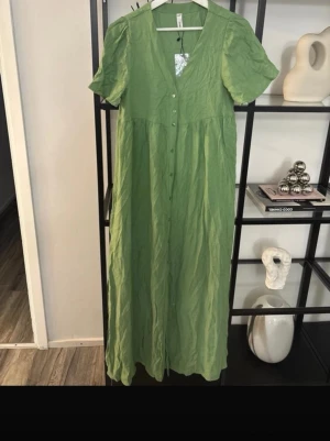 Grön långklänning med knappar - Lång grön klänning med korta ärmar och v-ringning. Klänningen har knappar framtill och är tillverkad i ett lätt, luftigt material som passar perfekt för varmare dagar. Enkel och stilren design med avslappnad passform.