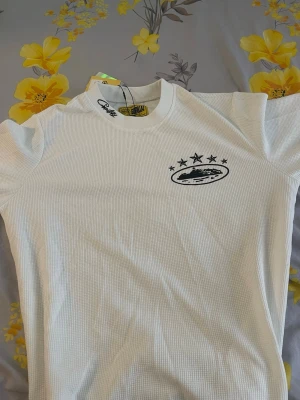 Vit kortärmad t-shirt från Ripndip - Vit kortärmad t-shirt från Ripndip med svart tryck på bröstet som visar ett motiv med stjärnor och logga. T-shirten har en ribbad struktur och rund halsringning. Perfekt för en chill och stilren look.