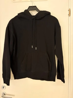 Svart basic hoodie med ficka - Svart hoodie med luva och ficka på magen. Det finns snören med silvriga stopp i slutet av snöret. 