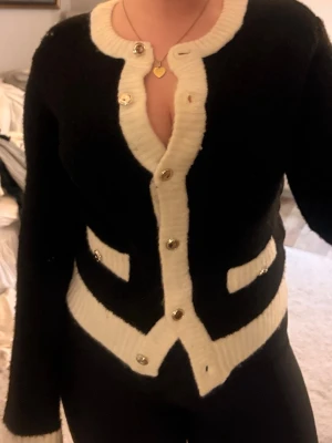 Svart kofta med vita detaljer - Säljer en svart kofta med vita kanter och dekorativa fickor. Koftan har guldknappar framtill och är tillverkad i ett mjukt, stickat material. Långärmad och med en klassisk, lite lyxig look.🤑🥰