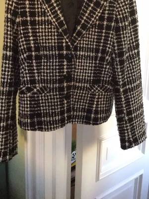 Svartvit rutig kavaj med knappar - Stilren svartvit kavaj med rutigt mönster och tre knappar framtill. Kavajen har klassisk krage, raka ärmar och två fickor fram. Materialet ser ut att vara tjockt och strukturerat, perfekt för en trendig look. Passar till många olika outfits.