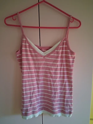 Rosa och vit randig linnetopp - Söt rosa och vit randig linnetopp med smala axelband och v-ringad hals. Toppen har ribbade kanter vid halsringning och nederkant, perfekt för varma dagar. Passar till jeans eller kjol för en avslappnad look.