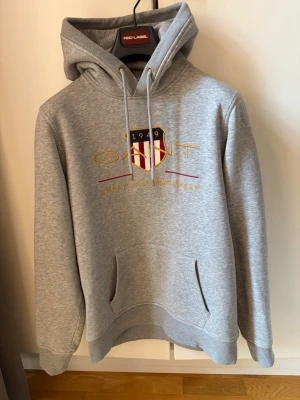 Gant Shield Hoodie - Snygg grå hoodie från Gant med broderad logga och sköld på bröstet. Hoodien har dragsko i huvan och en stor magficka. Tillverkad i mjukt bomullsmaterial och har ribbade muddar vid ärmar och nederkant. Perfekt för en avslappnad och sportig stil. 🎉Hoodien köpt på Boozt för 689kr på rea från 1400kr. 🎉För mer info skriv på dm!
