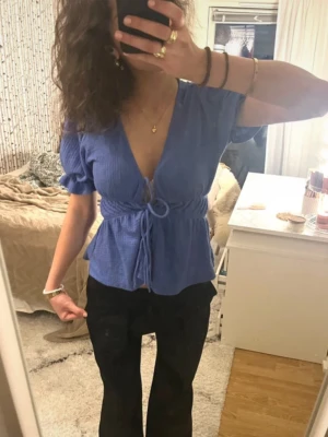 Blå omlottopp med knyt i midjan - Supersöt blå omlottopp med djup v-ringning och knyt i midjan. Toppen har korta ärmar med volangdetalj och en lätt ribbad struktur. Köpt på Gina. Perfekt nu till våren/sommaren💗
