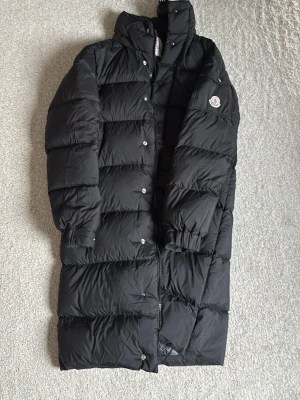 Svart lång dunjacka från Moncler - Snygg svart lång dunjacka från Moncler med hög krage och dragkedja samt tryckknappar. Jackan har Moncler-logga på ärmen och är fylld med dun för extra värme. Perfekt för kalla vinterdagar och har en stilren, puffig design.