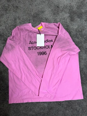 Rosa långärmad topp Acne Studios - Cool rosa långärmad topp från Acne Studios med svart text 'STOCKHOLM 1996' framtill. Oversized fit och rund halsringning. Perfekt för dig som gillar streetwear och vill sticka ut med en färgstark look.