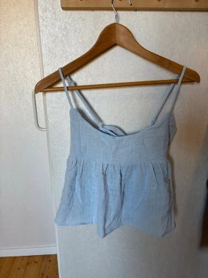 Ljusblå linnetopp med rosett - Supersöt ljusblå linnetopp med tunna axelband och en stor rosett i ryggen. Toppen har en luftig och flowy passform, perfekt för varma dagar. Materialet känns svalt och lätt mot huden.