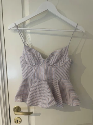 Randig peplum linnetopp med smala band - Snygg randig linnetopp med peplumskärning och smala axelband. Toppen har kupor och tunna blå, orangea och vita ränder som ger en fräsch sommarkänsla. Perfekt till jeans eller kjol för en trendig look. Storlek xs