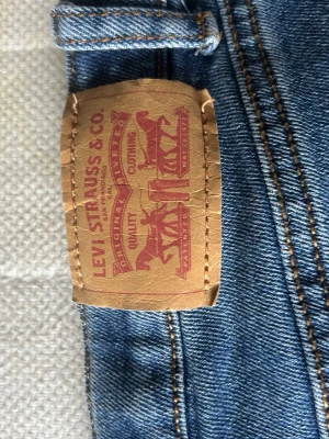 Blå Levi's jeans med klassisk söm - Ett par klassiska blå jeans från Levi's med den ikoniska läderpatchen bak i midjan och snygga kontrastsömmar. Jeansen har den typiska V-formade sömmen på bakfickan och är tillverkade i slitstark denim. Perfekta för en avslappnad och tidlös stil.