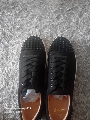 Svarta sneakers med nitar från Louboutin - Svarta sneakers med coola svarta nitar på tån, klassiska svarta skosnören och vit sula. Yttermaterialet är tyg och sulan är röd undertill, vilket är typiskt för Louboutin. Perfekta för dig som vill sticka ut med din stil.
