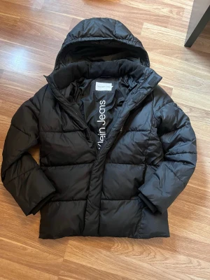Svart pufferjacka från Calvin Klein - Snygg svart pufferjacka från Calvin Klein Jeans med huva och diskret logga på insidan. Jackan har quiltad design, dragkedja framtill och är tillverkad i ett glansigt, vattenavvisande material. Perfekt för kalla vinterdagar och ger en stilren look. Nypris 1999 kr, köpt på Kids brand store.